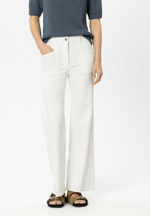 Person trägt weiße, hoch taillierte, weit geschnittene Hosen, ein blaues kurzärmliges Strickoberteil und braune Sandalen mit Schnallen, steht mit einer Hand in der Tasche.