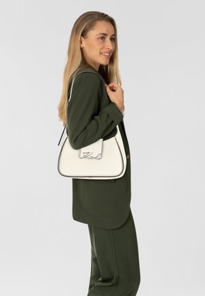 Witte leren handtas met zwarte bies en een kenmerkend logo, driehoekige vorm en een enkele schouderriem. Gedragen bij een donkergroene outfit.