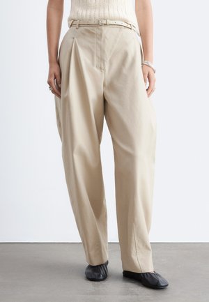 Pantalon beige taille haute à jambes larges avec ceinture assortie, porté avec des chaussures plates noires, montré de la taille aux pieds sur fond uni.