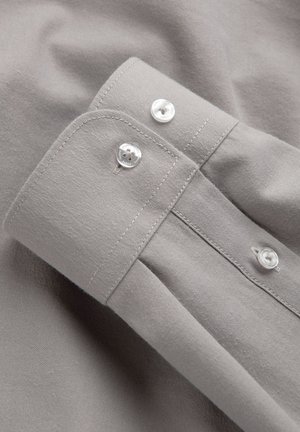 Poignet de chemise gris clair avec deux boutons blancs et coutures visibles, posé à plat sur un fond en tissu assorti.