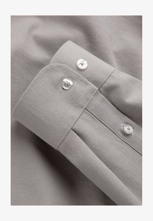 Poignet de chemise gris clair avec deux boutons blancs et coutures visibles, posé à plat sur un fond en tissu assorti.