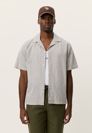 Kurzärmeliges Hemd mit Knöpfen und dünnen vertikalen Streifen in Grau und Weiß. Wird über einem weißen Tanktop getragen und mit grünen Hosen kombiniert.