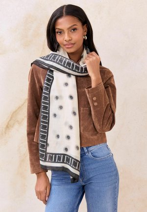 Ľahký šál s vzorom v bielej farbe s čiernymi bodkami, ohraničený geometrickým dizajnom. Nosený cez hnedú semišovú bundu a riflovinu.