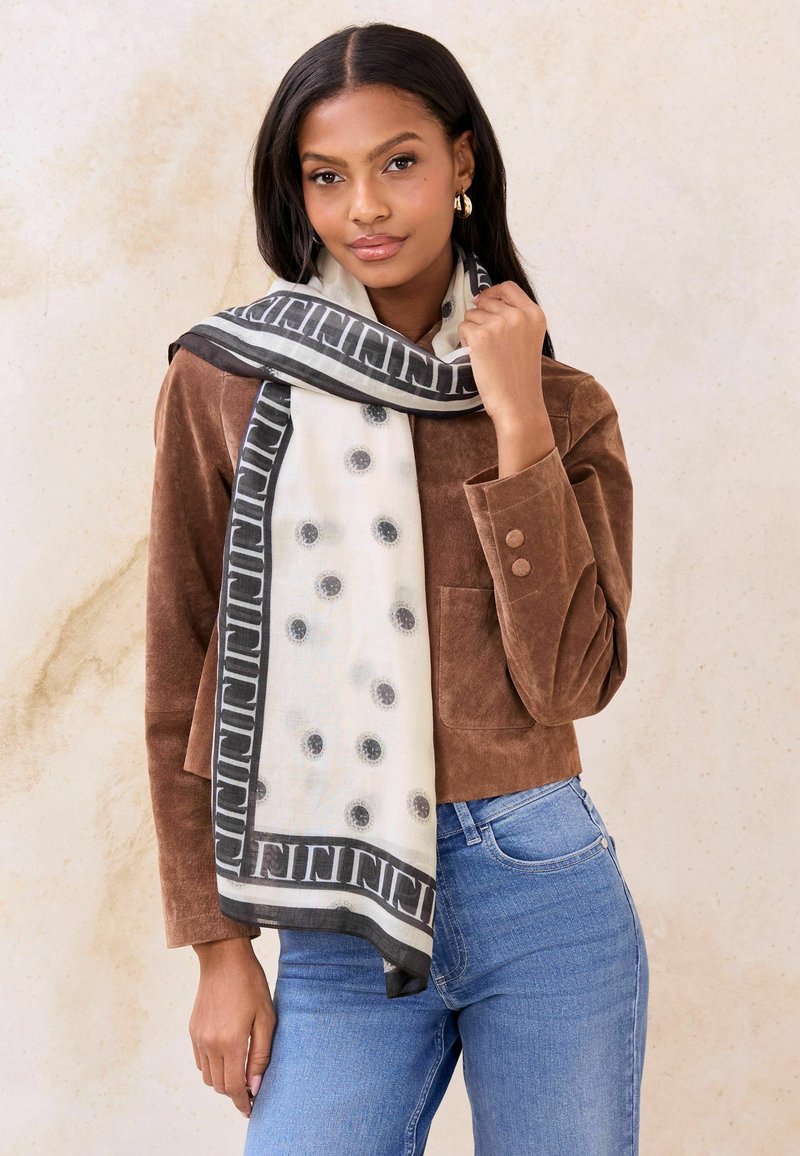 Lagani šal bijele boje s crnim točkama, obrubljen geometrijskim uzorkom. Nošen preko smeđe jakne od antilopa i trapera.