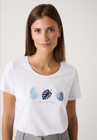 Junge Frau mit glattem braunem Haar trägt ein weißes Shirt mit blau- und schwarzbedruckten Blättern über dem Text "NUR Gemütlichkeit", kombiniert mit grauen Hosen.