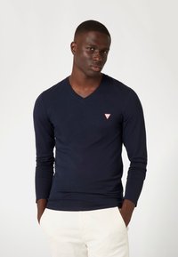 Navy lange mouwen V-hals shirt met een getailleerd ontwerp. Heeft een klein rood driehoekig logo op de borst. Soepele textuur, katoenmix materiaal.