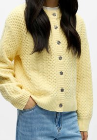 Keltainen neulottu neuletakki, jossa on tekstuurikuvio ja resorit hihansuissa, hopeiset napit kiinnityksessä ja pyöreä kaula-aukko. Yhdistetty sinisiin farkkuihin.