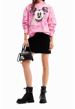 Persona vistiendo una sudadera con capucha rosa de Mickey Mouse, una falda corta negra, botas negras con cordones y sosteniendo un bolso negro con tachuelas.