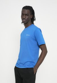 Μπλε βαμβακερό μπλουζάκι με κοντά μανίκια, στρογγυλή λαιμόκοψη και μικρό λευκό λογότυπο στο στήθος. Λείο ύφασμα και κανονική εφαρμογή.