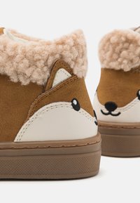 Brune semskede sko med en fluffy beige krage, med et design af en hvid og sort rævansigt på hælen og en tekstureret gummisål.