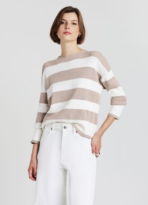 Giovane donna con capelli corti e castani che indossa un maglione a righe beige e bianco e pantaloni bianchi, in piedi con una mano in tasca.