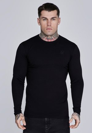 Homme avec des tatouages au cou et aux mains, portant une chemise noire unie à manches longues et un pantalon foncé, debout devant un fond gris.