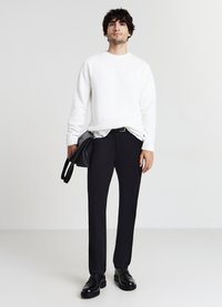 Weißer strukturierter Pullover, schwarze maßgeschneiderte Hose, schwarze Schuhe und eine schwarze Tasche. Der Pullover hat einen runden Halsausschnitt und das Outfit ist lässig.