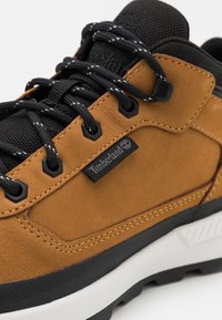 Baskets en cuir marron avec des accents en tissu noir, lacets noirs, œillets renforcés et semelle en caoutchouc blanche. Logo Timberland visible.