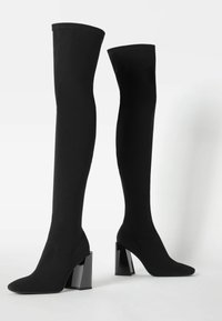 Botas negras por encima de la rodilla hechas de un tejido elástico, con punta afilada y grandes tacones de bloque con un acabado brillante.