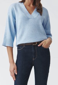 Jasnoniebieski sweter w serek z miękkiego materiału, z rękawami 3/4 i ściągaczami na rękawach. W połączeniu z ciemnoniebieskimi dżinsami i brązowym paskiem.