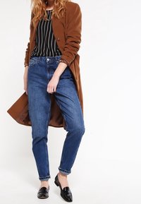 Cappotto lungo marrone su una maglietta a righe bianche e nere, abbinato a jeans azzurri chiari e mocassini neri, creando un outfit casual a strati.
