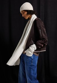 Giubbotto bomber marrone con polsini a coste, sciarpa di maglia grigio chiaro, berretto crema e jeans a gamba larga blu. I contrasti di texture sono evidenti nei tessuti.