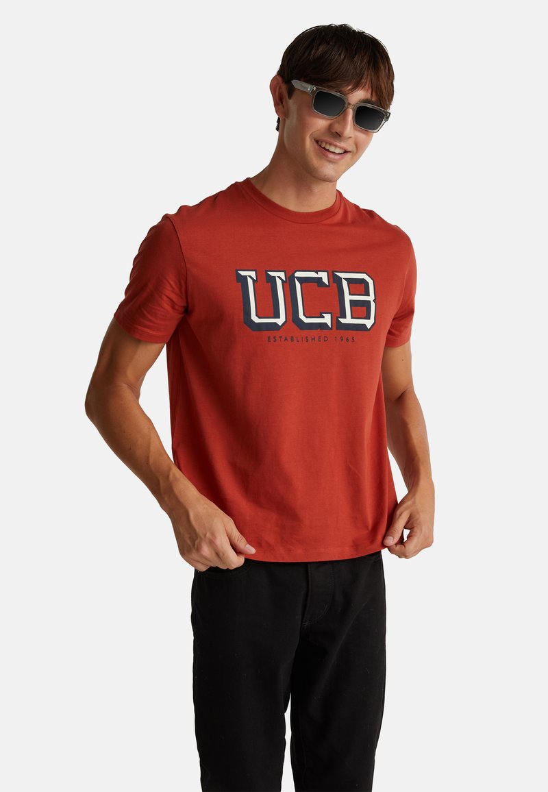 Joven sonriendo, lleva gafas de sol de montura transparente, una camiseta roja de UCB y pantalones negros, sujetando el dobladillo de la camiseta con ambas manos.