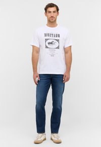 Biała koszulka z krótkim rękawem, ozdobiona czarnym grafikiem z napisem "MUSTANG" i obrazkiem koni, zestawiona z niebieskimi wąskimi dżinsami i beżowymi trampkami.