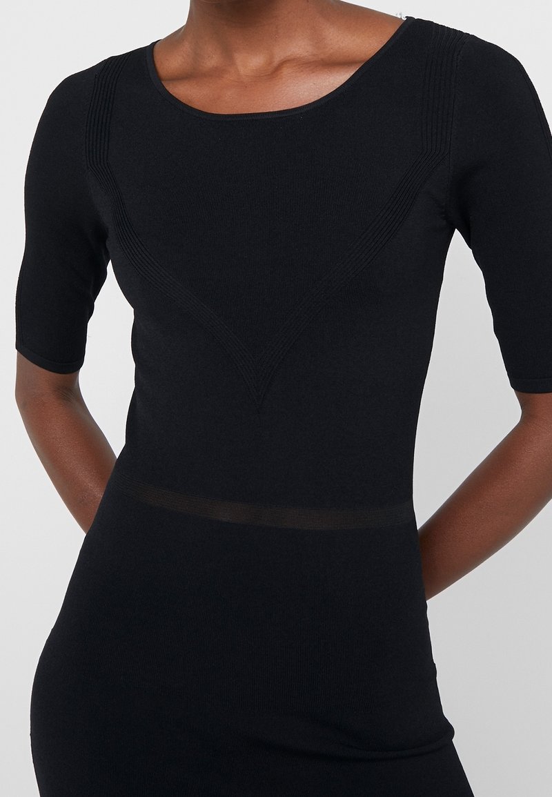 Femme portant une robe noire cintrée avec des détails texturés en forme de V sur la poitrine et des manches mi-longues, debout devant un fond clair uni.