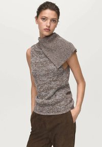 Femme portant un haut en tricot sans manches marron et gris avec un col asymétrique, associé à un pantalon marron foncé, se tenant devant un fond uni.