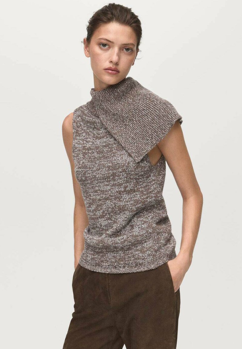 Femme portant un haut en tricot sans manches marron et gris avec un col asymétrique, associé à un pantalon marron foncé, se tenant devant un fond uni.