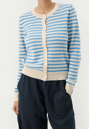 Femme portant un cardigan beige et bleu à rayures horizontales boutonné avec manches longues, rentré dans un pantalon bleu marine foncé avec poches.