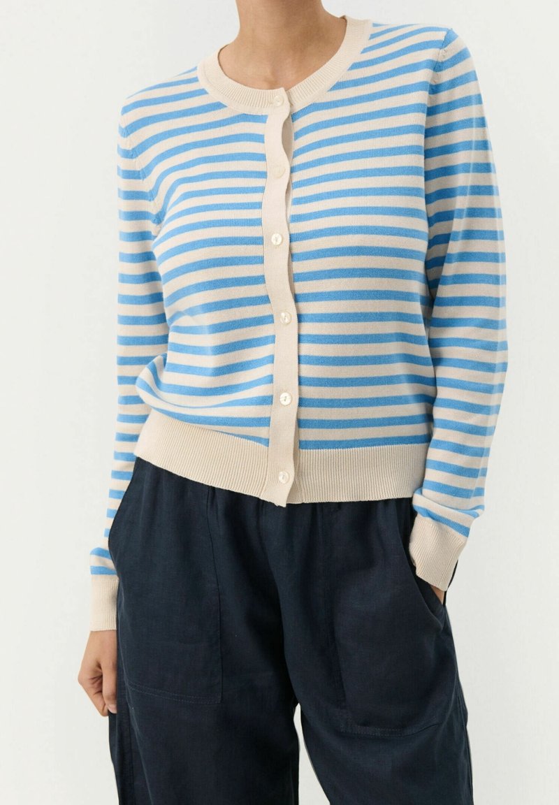 Femme portant un cardigan beige et bleu à rayures horizontales boutonné avec manches longues, rentré dans un pantalon bleu marine foncé avec poches.