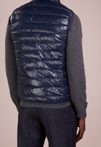 Muž v lesklé námořnické modré prošívané vestě přes šedý svetr a tmavé kostkované kalhoty stojící na jednobarevném pozadí.