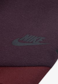 Mikina Nike s textúrovanou tkaninou v tmavo fialovej a marhuľovej farbe, s čiernym logom umiestneným na viditeľnom mieste.