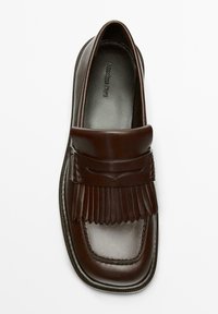 Mocassin en cuir marron avec une semelle plate, comportant un bout renforcé cousu et un détail à franges sur le cou-de-pied. Texture lisse et design élégant.