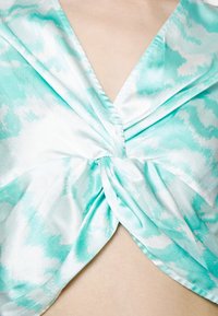 Turkusowa satynowa bluzka z węzłem z przodu i delikatnym, chmurowym wzorem. Gładka faktura, dekolt w kształcie V i zwisający materiał.