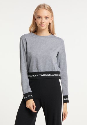 Junge Frau trägt ein graues Langarmshirt mit schwarzen Bündchen und Saum mit weißem ‚ATHLSR‘-Schriftzug, kombiniert mit schwarzen Hosen mit weißen Streifen.