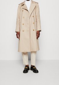 Béžový dvouřadý trenčkot se zlatými knoflíky, širokými klopami a nášivkami na ramenou. Kombinovaný s krémovými kalhotami a černými botami se zlatými doplňky.