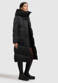 Schwarzer, gesteppter langer Steppmantel mit hohem Kragen, Reißverschluss und seitlichen Taschen. Kombiniert mit kniehohen schwarzen Stiefeln.