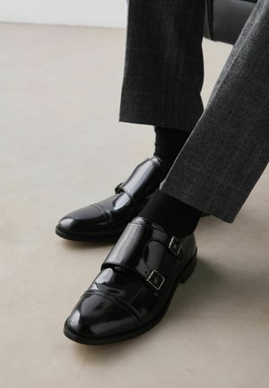 Black polished double monk strap shoes worn with dark gray trousers and black socks on a light floor.