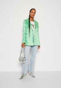 Sacou cu model de zebră verde, dintr-un material moale, asortat cu un tricou alb cu grafic, blugi deschiși evazați și cizme metalice argintii până la gleznă.