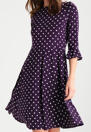 Donna che indossa un vestito viola al ginocchio con pois bianchi e maniche a tre quarti con polsini arruffati.