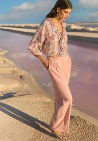 Floral, ruffled blouse in soft pink and blue tones; beige, tapered pants with pleats; metallic-accented heels. Natural landscape background.