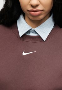 Μαρόν φούτερ με ριμπ λαιμόκοψη, με λευκό λογότυπο Nike. Φαίνεται ανοιχτό μπλε, υφασμάτινο πουκάμισο με γιακά από κάτω.