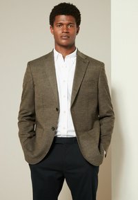 Blazer marrón texturizado con dos botones, solapas en pico y bolsillos laterales, llevado sobre una camisa blanca de botones y pantalones oscuros.