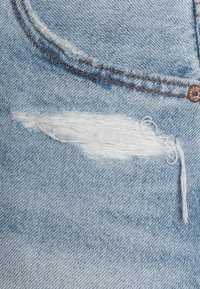 Světle modrá džínsovina s oprýskanou oblastí a třepenými okraji. Oranžové prošívání podél kapsy a na švu.