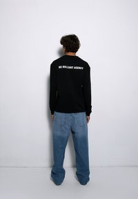 Czarna koszula z długim rękawem z białym napisem "NO BULLSHIT AGENCY" na plecach, w połączeniu z luźnymi jasnoniebieskimi dżinsami.