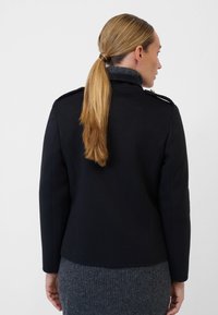 Black tailored jacket with a structured collar, featuring gold hardware details, set against a grey textured dress.