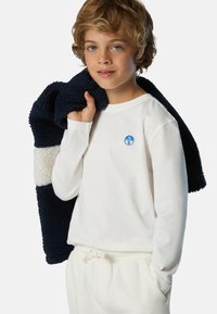 Camicia bianca a maniche lunghe con un distintivo blu, indossata con pantaloni jogger crema. Giacca in pile blu navy con tessuto textured appoggiata su una spalla.