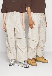 Beige cargo bukser med elastisk talje, har sidelommer og en løs pasform. Sammen med en brun topp og forskellige sneakers i sølv og gul.