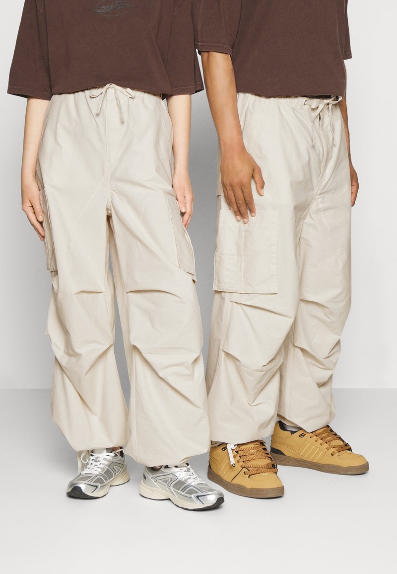 Beige cargo bukser med elastisk talje, har sidelommer og en løs pasform. Sammen med en brun topp og forskellige sneakers i sølv og gul.