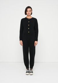 Μαύρο σετ από ριπ πλεκτό ύφασμα που περιλαμβάνει μακρυμάνικο τοπ με χρυσά κουμπιά και παντελόνι με στενή γραμμή, ολοκληρωμένο με μαύρα αθλητικά παπούτσια.