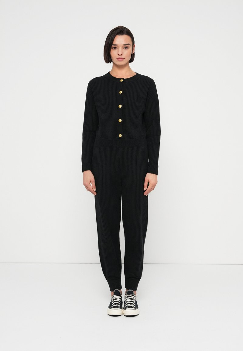 Μαύρο σετ από ριπ πλεκτό ύφασμα που περιλαμβάνει μακρυμάνικο τοπ με χρυσά κουμπιά και παντελόνι με στενή γραμμή, ολοκληρωμένο με μαύρα αθλητικά παπούτσια.